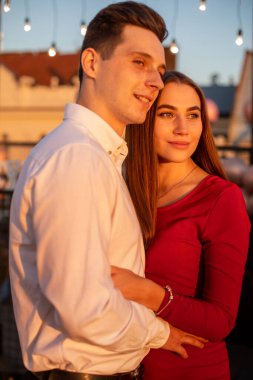 Bir çift, bir akşam toplantısında birbirlerine yakın dururken bir anı paylaşır. Gökyüzü günbatımı renkleriyle parlıyor ve sicim ışıkları yanıyor.