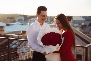 Adam gün batımında çatıda bir kadına büyük bir buket kırmızı gül veriyor. Arka planda şehir binaları var..