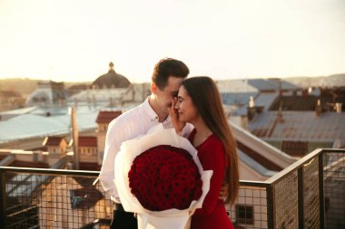 İki insan gün batımında çatıda büyük bir buket kırmızı gül tutarken birbirlerine gülümserler..