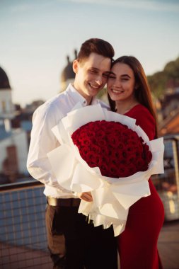 Bir çift çatıda birlikte duruyor, büyük bir buket kırmızı gül tutuyorlar. Gülümsüyorlar ve şehirdeki akşam atmosferinin tadını çıkarıyorlar..