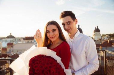 Bir kadın bir buket kırmızı gül tutarken bir adam arka planda güneş batarken onun yanında duruyor..