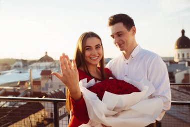 Bir çift bir buket gülle çatıda duruyor. Kadın gülümseyerek yüzüğünü gösteriyor. Güneş şehrin üzerinde batıyor ufuk çizgisi.