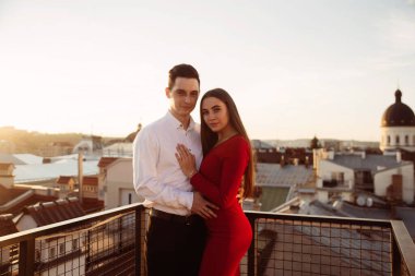 Bir çift, güneş şehrin üzerinde batarken çatıda yakın poz veriyor. Manzaradan ve birbirlerine eşlik etmekten hoşlanırlar..