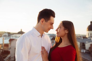 İki insan gün batımında çatıda birbirlerinin gözlerine bakar. Arka planda binalar ve su vardır.