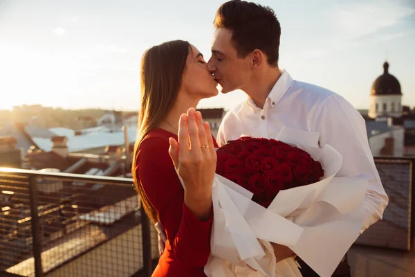 Gün batımında çatıda iki kişi öpüşür. Biri büyük bir buket kırmızı gül tutuyor ve nişan yüzüğü gösteriyor..