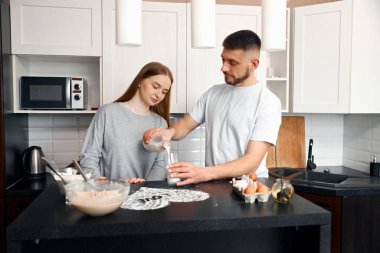 İki kişi modern bir mutfakta kahvaltı hazırlıyorlar. Biri süt dökerken diğeri izliyor. Yumurta ve kase gibi malzemeler tezgahta..