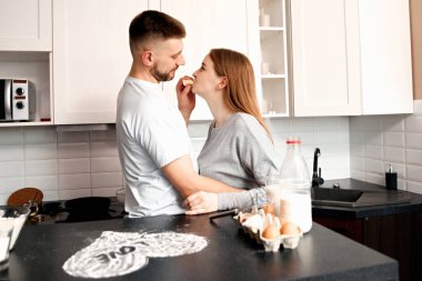 Biri diğerini beslerken, bir çift mutfakta durup hafif bir anı paylaşıyor. Mutfak modern ve aydınlık. Etrafında yiyecek parçaları var..