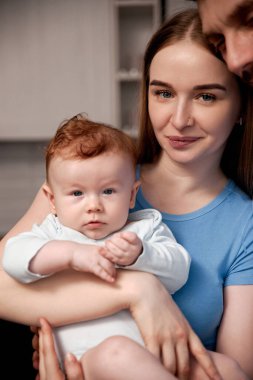 Ebeveynler bebeklerini bir evde tutarken gülümserler. Bebeğin kıvırcık kızıl saçları var ve meraklı görünüyor. Sade mobilyalar ve sıcak renklerle ortam aydınlık..