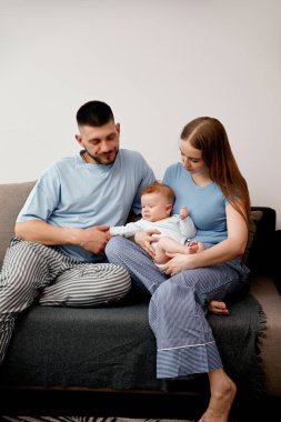 Bir anne ve baba oturma odasındaki bir kanepede rahatlar. Açık renk giyinmiş bebeklerini kucaklıyorlar..