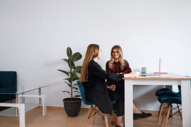 İki kadın, ofis odasında bir masada, bitkiler, dizüstü bilgisayarlar ve ofis mobilyalarıyla sohbet ediyorlar..