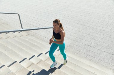 Canlı aktif giyimli atletik kadın açık havada merdivenlerden yukarı koşuyor, spor ve sağlığa bağlılığını gösteriyor, güneş ışığı desenli betonu aydınlatıyor, dinamik bir atmosfer yaratıyor..