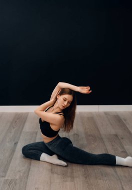 Aktif kıyafetli genç bir kadın spor salonunda yerde otururken vücudunu esnetiyor. Egzersiz rutinine odaklanırken gülümsüyor..