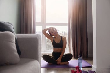 Bir kadın yoga minderinde oturur ve sabahları evde ağırlık çalışırken boynunu uzatır..