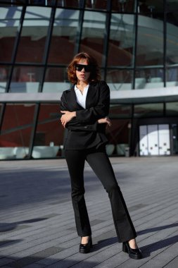 Şık, siyah takım elbiseli, modern ofis binasının yanında duran zarif bir iş kadını..