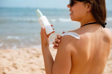 Plajda güneş kremi ve güneş kremi şişesi tutan çekici genç bir kadın. Cilt bakımı, güneş koruması ve yaz yaşam tarzı kavramı. Deniz kenarında tatil yaparken cilt bakımı ve ultraviyole ışınlardan korunmak için losyon kullanan bir kadın..