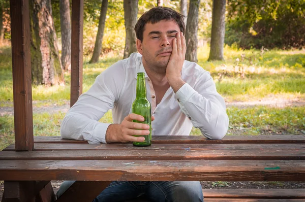Részeg ember ül egy padon, és a gazdaság egy sörösüveg — Stock Fotó
