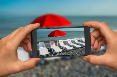 Taking photo of sunbathing beds and red umbrella with phone clipart