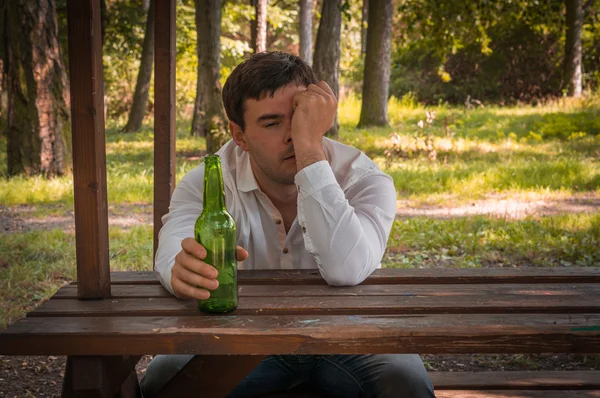 Részeg ember ül egy padon, és a gazdaság egy sörösüveg — Stock Fotó