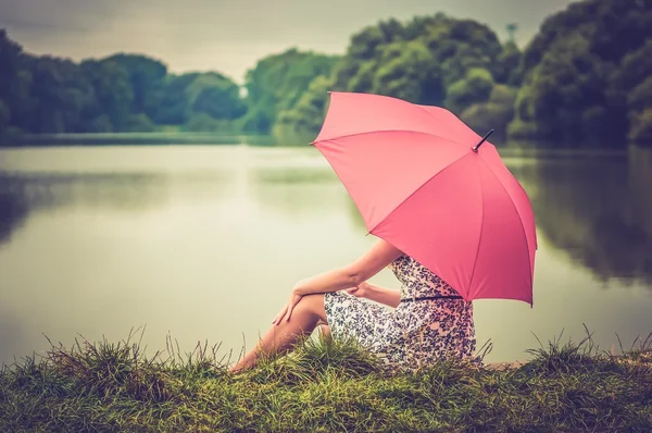 Junge schöne Frau mit rotem Regenschirm am Teich sitzend - — Stockfoto