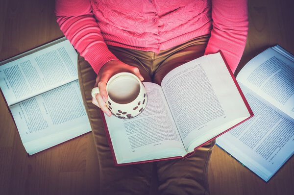 Женщина держит в руках чашку кофе, сидит на полу и учится по открытой книге
