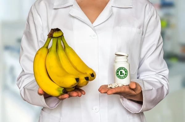 Joven médico sosteniendo plátanos frescos y frasco de pastillas con vitaminas y compararlos — Foto de Stock