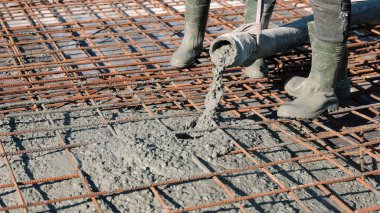 Concrete being poured over steel reinforcement mesh. Construction workers in boots pouring wet concrete onto a rebar grid, preparing the foundation slab for a building structure