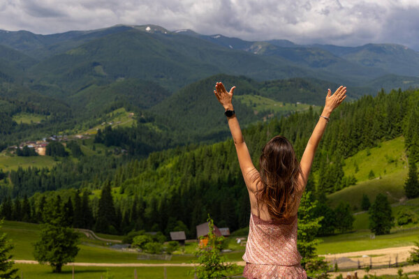 Woman celebrating freedom in mountain valley view. A woman raises her arms in joy while enjoying a panoramic view of lush green mountains and a peaceful rural landscape