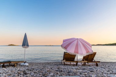 Şemsiyeli ve şezlonglu romantik plaj günbatımı. Gün batımında, iki ahşap şezlonglu, romantik ve rahatlatıcı tatil temaları için mükemmel bir yer.