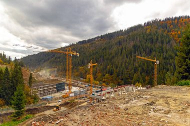 Dağ inşaatında birden fazla vinç var. Dağ vadisinde büyük bir inşaat alanı. Birkaç sarı kule vinci, temeller ve bulutlu gökyüzünün altında orman yamaçları var..