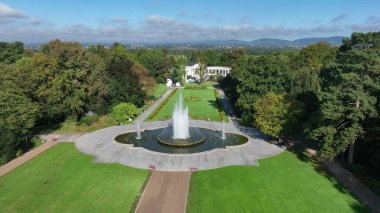 Büyük çeşmeli kamu parkının havadan görünüşü. İnsansız hava aracı büyük bir çeşme, yürüyüş yolları, ağaçlar ve uzak tepelerin yer aldığı yeşil şehir parkının manzaralı bir manzarasını yakalar.