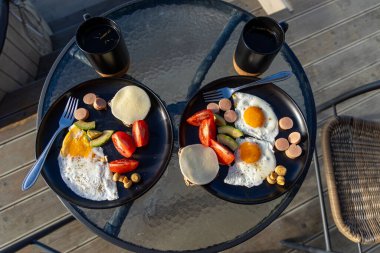 Yumurta ve kahveyle birlikte sağlıklı bir açık hava kahvaltısı. İki tabak sahanda yumurta, sebze, sosis, peynir ve kahve teras masasında huzurlu bir dağ sabahına hazır.