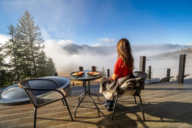 Kadın kahvaltı yaparken dağ manzarasına hayran kalıyor. Bir kadın terasta oturuyor, battaniyeye sarılmış, kahvaltısını yapıyor ve huzur içinde dağların sisli manzarasını seyrediyor.