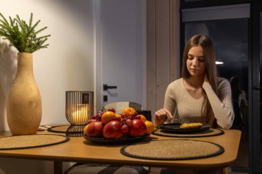 Genç kız evde tek başına yemek yiyor. Genç kız gece rahat bir yemek masasında oturuyor, yanında taze meyve ve mum ışığı olan bir krep yiyor.
