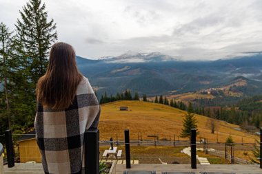 Terastan dağ manzarasına bakan bir kadın. Ekoseli battaniyeye sarılı bir kadının, tahta bir güvertede durup sisli bir dağ vadisine hayran olduğu arka plan görüntüsü.