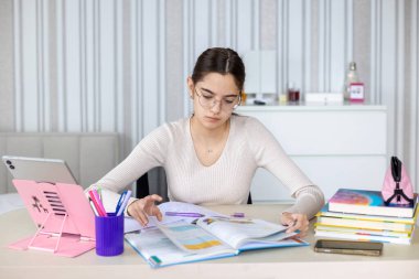 Evde ders çalışan odaklı bir genç kız. Okul malzemeleri, kitaplar ve tabletle çevrili, ders kitabı okuyan ve evde ders notları yazan genç bir kız.