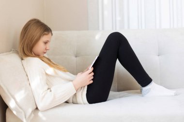 Genç kız akıllı telefon kullanarak kanepede dinleniyor. Hafif bir kanepede uzanan, sakin ve rahat bir ortamda akıllı telefonuna odaklanmış genç bir kızın yan görüntüsü.