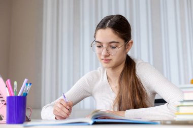 Masadaki not defterine çalışan ve yazan genç bir kız. Gözlüklü ve hafif bir kazak giyen odaklanmış genç bir kız, bir masada oturuyor ve evde ders çalışırken bir deftere yazıyor.