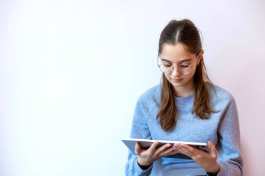 Gözlüklü genç kız beyaz arka planda tablet kullanıyor. Gözlüklü ve gri kazaklı odaklanmış genç bir kız, düz beyaz bir duvara yaslanırken tablet bir cihaz tutuyor ve kullanıyor.