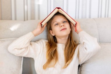 Kapalı mekanlarda kitap tutan oyuncu kız. Kanepede oturan genç bir kız başının üzerinde bir kitap tutulmuş, neşeli bir şekilde gülümsüyor.