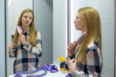 Genç kız aynada uzun saçlarını tarıyor. Genç bir kız, yansımasına bakarken uzun sarı saçlarını tarar ve evde sakin bir sabah bakım rutini gösterir.