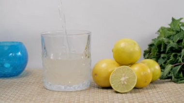 İçi ferahlatıcı limon suyuyla dolu bardağın içine su dökülüyor. Dilimlenmiş limon ve taze nane yaprağıyla servis edilen birkaç limonun yakından çekimi.