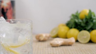 Büyük buz küpleri limon dilimli bir bardak taze soda / tonik suya düşüyor. Birkaç zencefilli ve dilimlenmiş limonlu jüt hasırının üzerinde nane yapraklarıyla saklanmış.