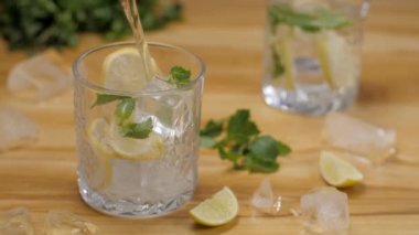 Dilimlenmiş limon ve nane yapraklı bir bardağa taze limon suyu dökülüyor. İki dilim limon ve ahşap bir masada bir bardak limon suyuyla birkaç buz.