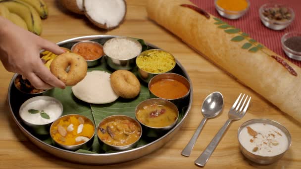 雌性手将梅杜 瓦达和其他南印度人的食物放在盘子里 一束香蕉 椰子半边 素食节大理和帕瓦萨姆碗放在一张木制桌子上 — 图库视频影像