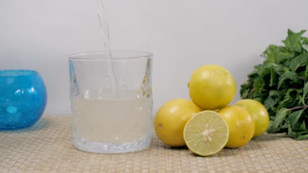 ガラスの半分の中に水が落ち さわやかなレモン汁で満たされます スライスされたレモンと新鮮なミントの葉で保持されたいくつかの全レモンのクローズアップショットは ジュートマットに残します — ストック動画