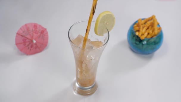 通気性の高い飲み物は 上にレモンスライスと透明なガラスに注ぎました 白い背景を背景に おいしいクリスピーなスナックで保管された小さなピンクの傘の閉鎖ショット — ストック動画