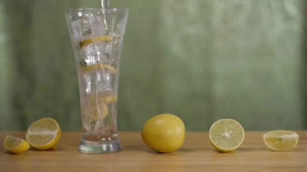 新鮮なレモン汁をアイスキューブで透明なガラスに注ぎます 緑の背景に木製のテーブルの上に一緒に保持全体のショットとスライスレモン — ストック動画