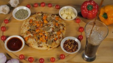 Bir bardak soğuk içecek lezzetli tavuk sosisli pizzayla servis ediliyor. Tahta bir masanın üzerinde kekik, peynir küpleri ve kırmızı biberli lezzetli vejetaryen olmayan bir pizzanın yakın çekimi.