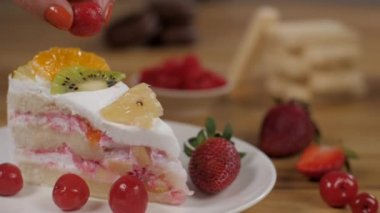 Bayan şef bir dilim kremalı meyveli kekin üzerinde sulu kırmızı bir kiraz tutuyor. Taze meyve, kivi, portakal, ananas, çilek ve kiraz gibi taze meyvelerle süslenmiş sünger sütlü pasta dilimi.