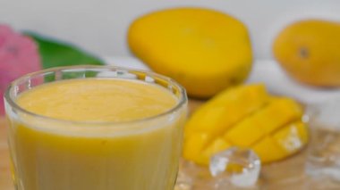 Şeffaf bir bardak taze mango milkshake ve meyve suyuna damlacıklar düşüyor. Dilimlenmiş sarı sulu mango ve beyaz arka planda bir arada tutulan birkaç buz küpünün yakın plan çekimi. 